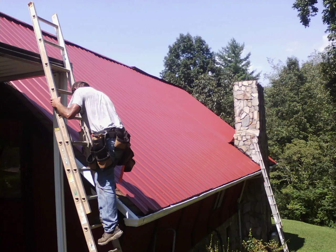 Roofer on a ladder beside a red metal roof and stone chimney — Soffit & Fascia Repair by Roofing Brothers in Napoleon